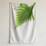 Un drapeau au dessin de fougère sur fond blanc suspendu grâce à es oeillets et des clous sur un mur texturé blanc. 