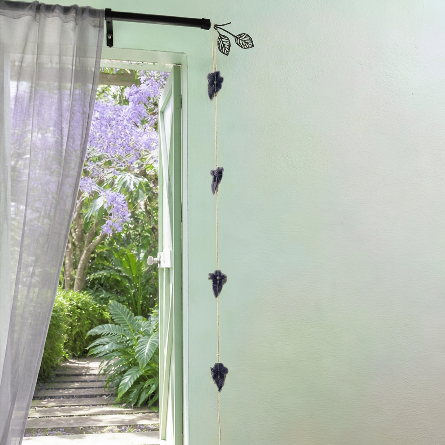 Guirlande décorative de feuilles sombres suspendue près d’une fenêtre ouverte sur un jardin tropical réunionnais en fleurs.